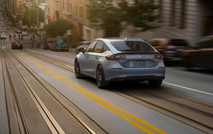 Rear driver-side view of the 2023 Honda Civic Sport Hatchback in Sonic Gray Pearl, driving on a city street.