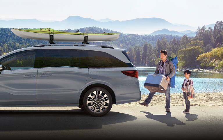 2027 Honda Odyssey Elite in Sonic Gray Pearl profile parked by a scenic lake with a family loading gear (Minivan).