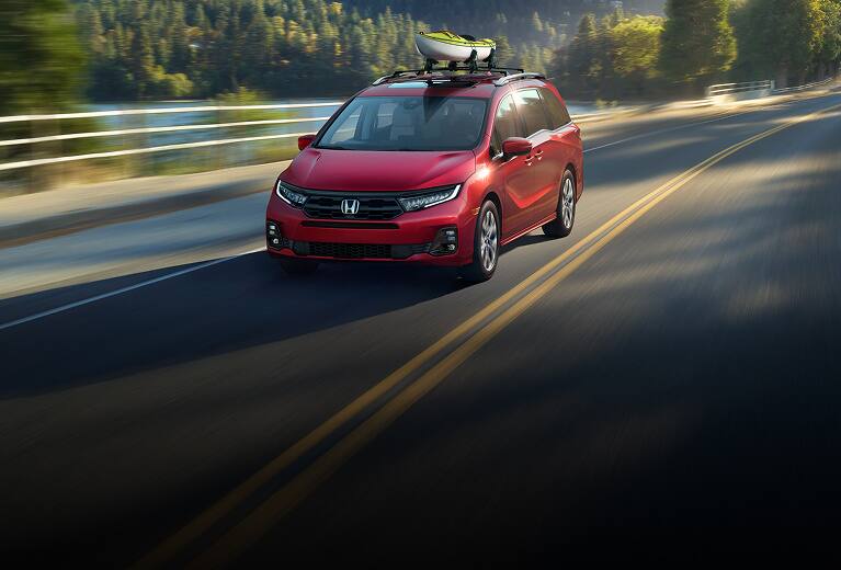 2027 Honda Odyssey Elite in Radiant Red Metallic II front driving on a scenic road near a lake and forest (Minivan).