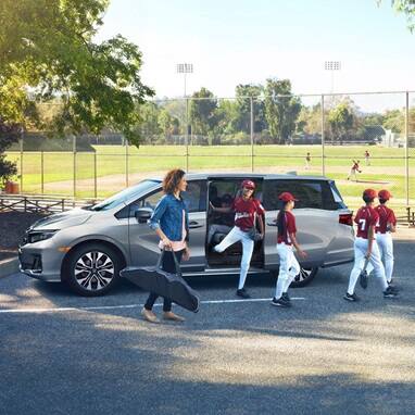 2027 Honda Odyssey Elite in Solar Silver Metallic parked at a baseball field with children, showcasing a spacious minivan for active families (Minivan).